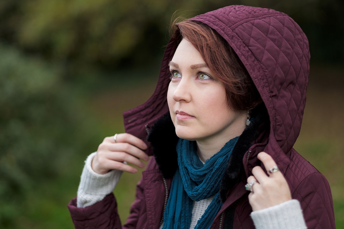 Portrait of a young woman with green eyes & red hair wearing a burgundy hood looking away from camera.
