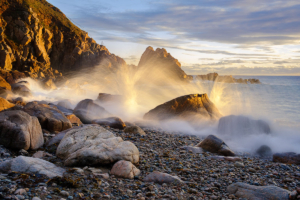 Waves crash onto Le Jaonnet Bay on the South coast of Guernsey, backlit by warm sunlight.