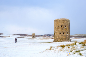 A man walks his dog across fresh snow near loophole towers on L'ancresse Common, Guernsey.