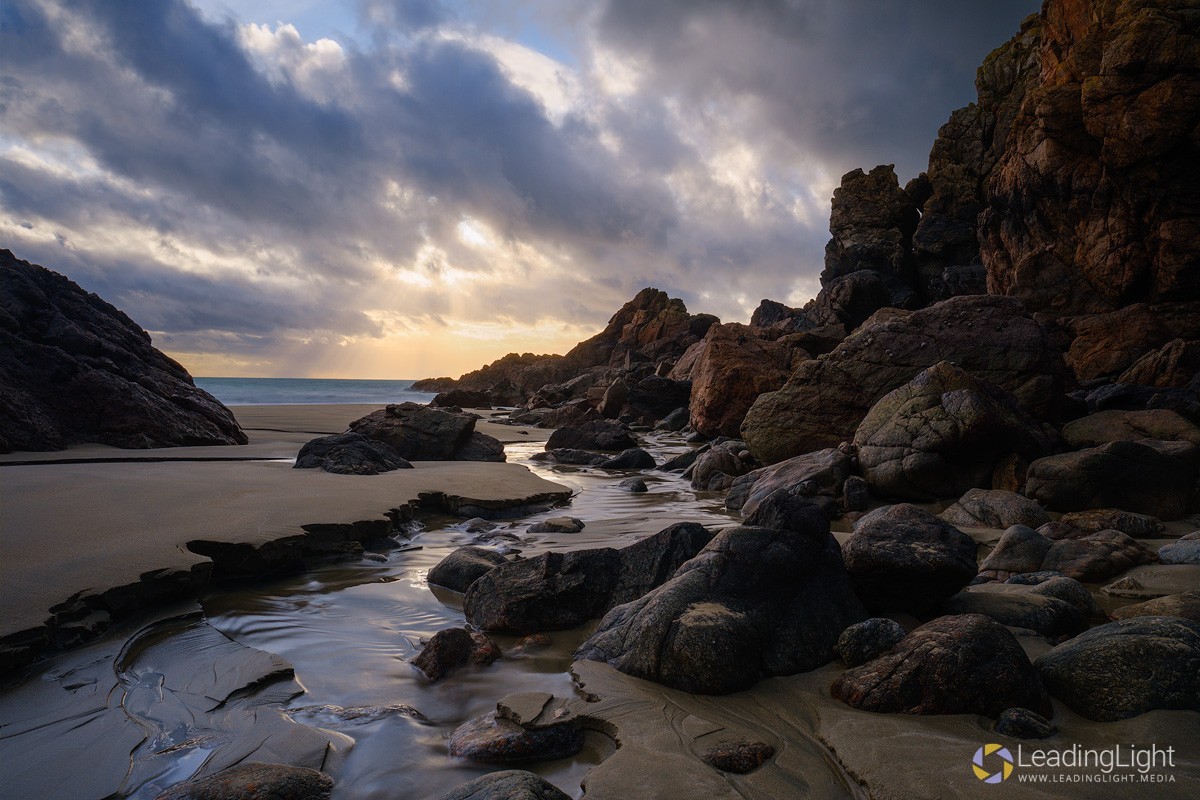 A stream, or rivulet, meanders around rocks in the sand at Le Jaonnet Bay, Guernsey at sunset